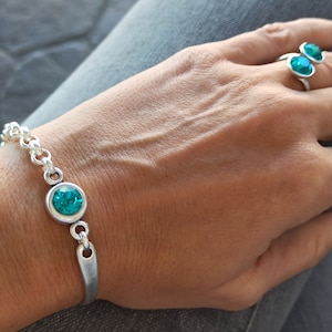 May include: A silver bracelet with a turquoise blue crystal charm. The bracelet is on a person's wrist. The person is also wearing a silver ring with two turquoise blue crystal stones.