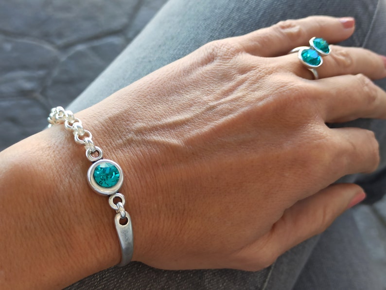 May include: A silver bracelet with a turquoise blue crystal charm. The bracelet is on a person's wrist. The person is also wearing a silver ring with two turquoise blue crystal stones.