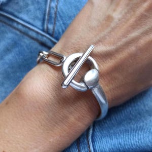 Può includere: Un braccialetto argentato con chiusura a toggle, con un anello circolare e una chiusura a barra. Il braccialetto è indossato su un polso, con uno sfondo in denim.