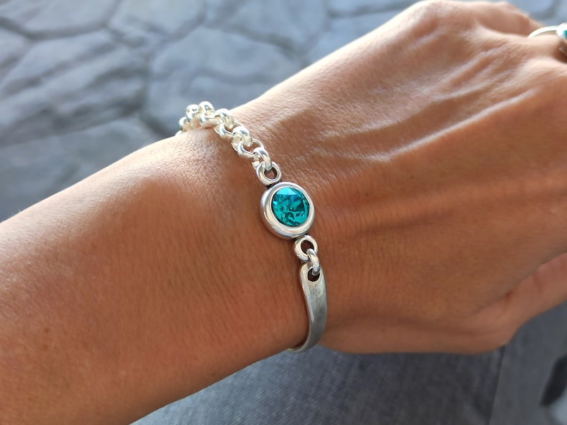 May include: A silver bracelet with a turquoise blue gemstone. The bracelet has a chain link design and a simple, curved bar closure.