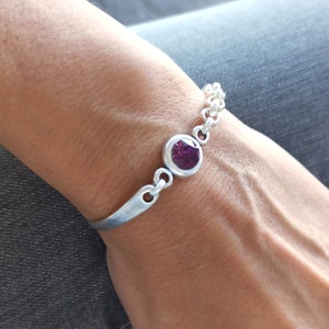 May include: A silver bracelet with a single purple gemstone set in a silver bezel. The bracelet has a chain link design and a simple, curved bar.