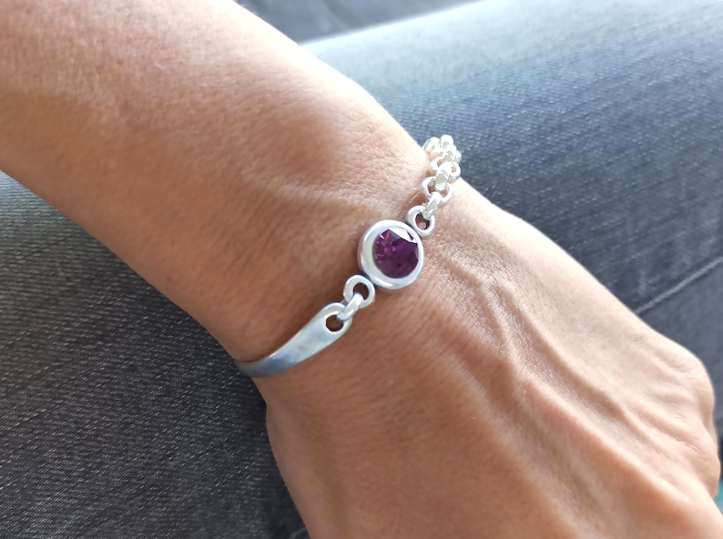 May include: A silver bracelet with a single purple gemstone set in a silver bezel. The bracelet has a chain link design and a simple, curved bar.