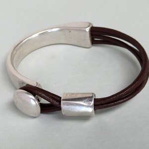 Puede incluir: Un brazalete de cuero marr&oacute;n con un cierre de metal plateado y una cuenta de metal plateado. El brazalete est&aacute; hecho de tres hebras de cuero.