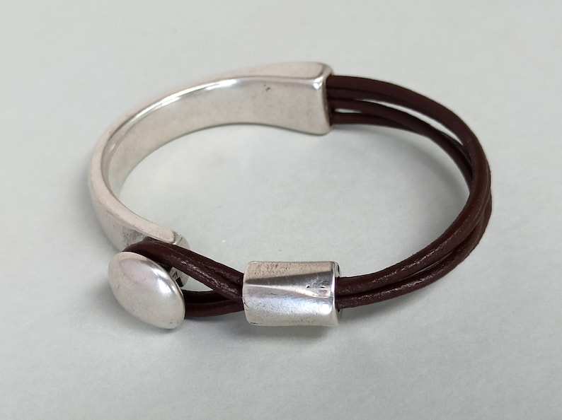 Puede incluir: Un brazalete de cuero marr&oacute;n con un cierre de metal plateado y una cuenta de metal plateado. El brazalete est&aacute; hecho de tres hebras de cuero.
