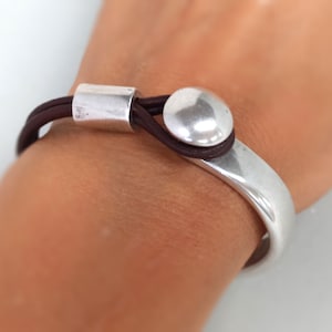 Puede incluir: Un brazalete de plata con una correa de cuero marr&oacute;n y un acento redondo de plata. El brazalete se lleva en la mu&ntilde;eca de una persona.
