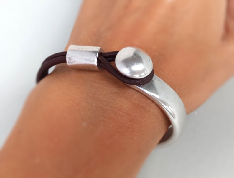 Puede incluir: Un brazalete de plata con una correa de cuero marr&oacute;n y un acento redondo de plata. El brazalete se lleva en la mu&ntilde;eca de una persona.