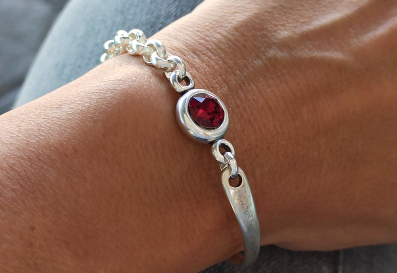 May include: A silver bracelet with a red gemstone. The bracelet has a chain link design and a simple, curved bar.