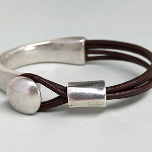 Puede incluir: Un brazalete de cuero marr&oacute;n con un cierre de metal plateado y dos cuentas de metal plateado. El brazalete est&aacute; hecho de tres hebras de cuero.