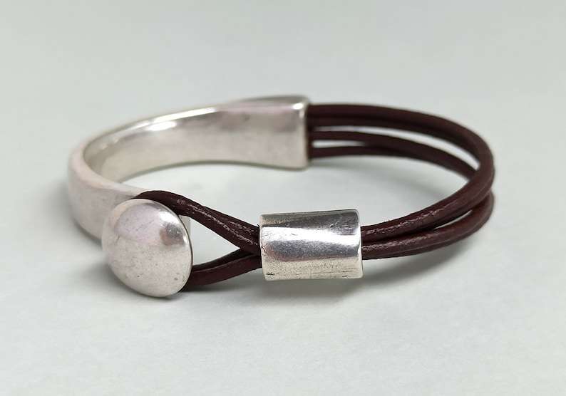 Puede incluir: Un brazalete de cuero marr&oacute;n con un cierre de metal plateado y dos cuentas de metal plateado. El brazalete est&aacute; hecho de tres hebras de cuero.