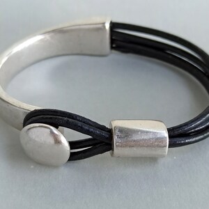 Puede incluir: Un brazalete de plata con una correa de cuero negro. La correa est&aacute; hecha de tres hebras de cuero y tiene un cierre de plata.