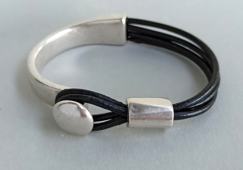 Puede incluir: Un brazalete de plata con una correa de cuero negro. La correa est&aacute; hecha de tres hebras de cuero y tiene un cierre de plata.