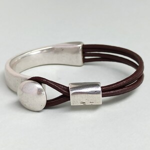 Puede incluir: Un brazalete de cuero marr&oacute;n con un brazalete de metal plateado y dos acentos de metal plateado. El brazalete est&aacute; hecho de tres hebras de cuero.