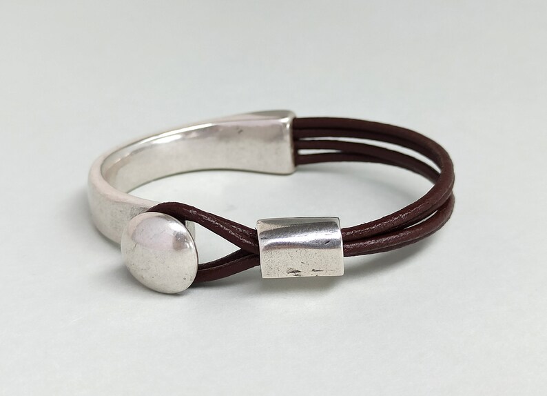 Puede incluir: Un brazalete de cuero marr&oacute;n con un brazalete de metal plateado y dos acentos de metal plateado. El brazalete est&aacute; hecho de tres hebras de cuero.
