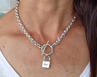 May include: A silver necklace with a thick chain. It has a toggle clasp and a small padlock pendant engraved with the word "love". The necklace is worn around the neck.