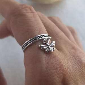 Può includere: Un anello d'argento con una fascia testurizzata e un ciondolo a forma di ape. L'anello è indossato su un dito.
