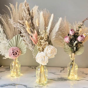 May include: Three decorative floral arrangements in glass bottles with fairy lights. Each arrangement features dried pampas grass, roses, and other dried flowers. The roses are in shades of pink and white, and the arrangements are set on a marble surface.