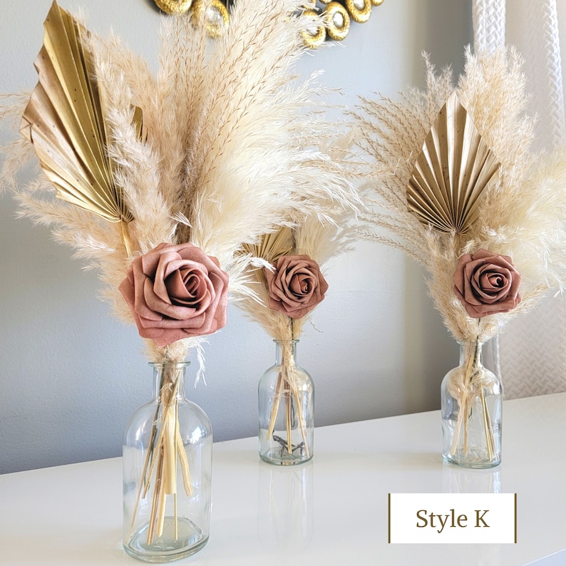 May include: Three glass vases filled with dried pampas grass and blush pink roses. The vases are on a white surface. The text "Style K" is in the lower right corner of the image.