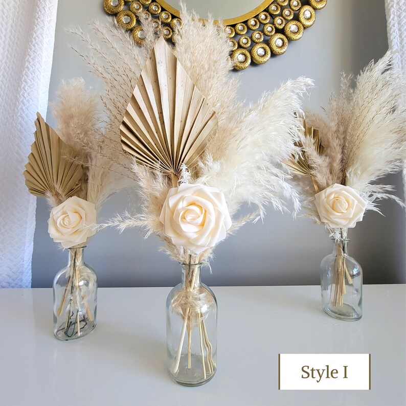May include: Three glass vases filled with dried pampas grass, dried palm leaves, and faux cream-colored roses. The vases are on a white surface with the text "Style I" below the center vase.