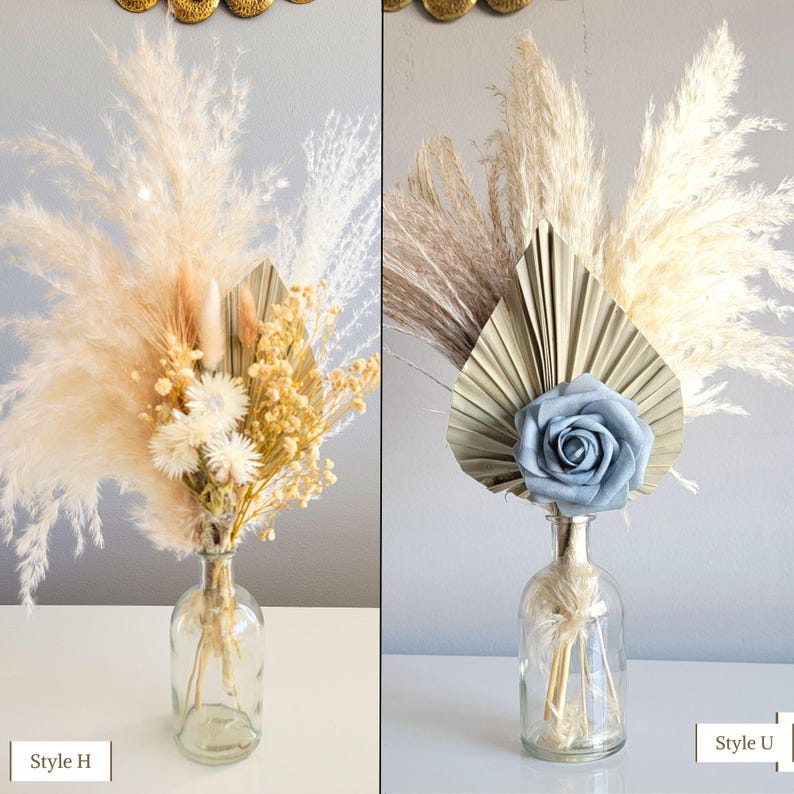 May include: Two dried flower arrangements in clear glass vases. The arrangement on the left features a mix of dried grasses, pampas grass, and small white flowers. The arrangement on the right features a large dried palm leaf, pampas grass, and a single blue silk rose.