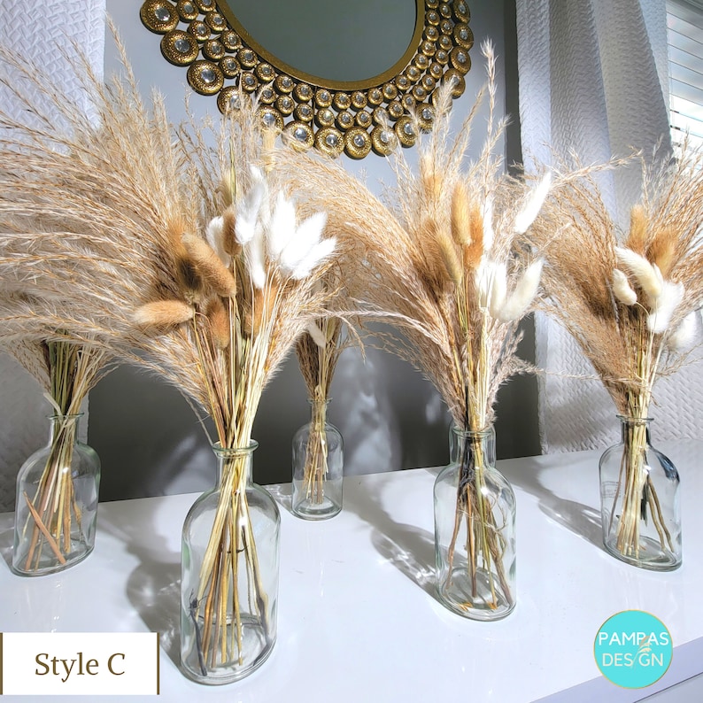 May include: Five clear glass vases filled with dried pampas grass and fluffy white bunny tails. The vases are arranged in a row on a white surface.