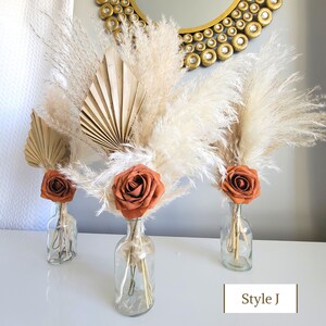 May include: Three glass vases with dried pampas grass, a dried palm leaf fan, and a single orange rose in each vase. The vases are on a white surface. The text "Style J" is in the lower right corner of the image.