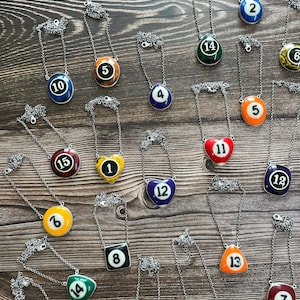 Peut inclure: Une collection de colliers avec des pendentifs en forme de boules de billard de différentes couleurs et numéros, incluant 1, 2, 4, 5, 6, 7, 8, 10, 11, 12, 13, 14 et 15. Les pendentifs sont fixés à de fines chaînes métalliques argentées, certains en forme de cœur ou de triangle.