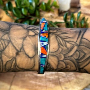 May include: A silver cuff bracelet featuring a central inlay of geometric segments in vibrant turquoise, bright orange, and iridescent white. The inlay is composed of small, angular pieces set flush within the polished metal frame of the bracelet.