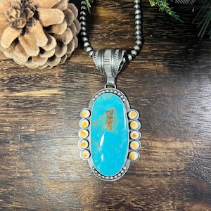 May include: A silver pendant necklace featuring a large, oval-shaped turquoise stone with brown matrix inclusions. The pendant is accented by ten small, round, cream-colored stones along the sides and hangs from a silver bead chain.