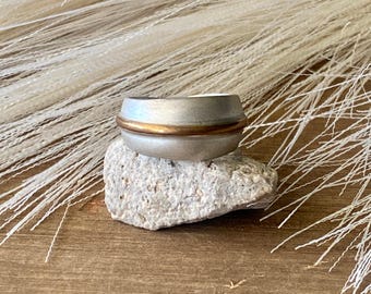Könnte beinhalten: Ein breiter Ring mit mattem, silberfarbenem Finish und einem dünnen, polierten, kupferfarbenen Mittelband. Der Ring hat ein leicht gewölbtes, konvexes Profil und eine glatte, gleichmäßige Textur auf der gesamten Metalloberfläche.