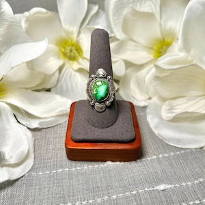 May include: A sterling silver ring featuring an oval-shaped green turquoise stone with dark matrix patterns. The setting is decorated with small silver beads and fan-shaped accents around the central stone, creating a detailed metal border.