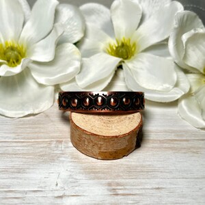 May include: A copper-toned metal cuff bracelet featuring a repeating pattern of raised circular studs and dark recessed geometric shapes. The band has a polished metallic finish along the top and bottom edges, showcasing a distinct contrast between the bright copper and the dark, textured center detailing.