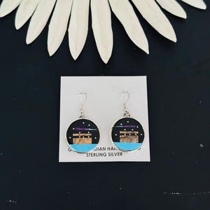 May include: Round sterling silver dangle earrings featuring a black inlay with a tan rectangular structure, a bright blue base, and small white dots representing stars. The earrings are attached to a white display card labeled STERLING SILVER.