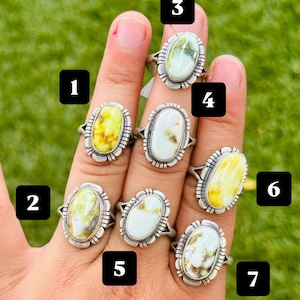 May include: Seven silver rings featuring oval-shaped stones in shades of white, yellow, and green, each labeled with a black square containing a white number from 1 to 7. The rings have decorative metal settings with etched line details around the edges.