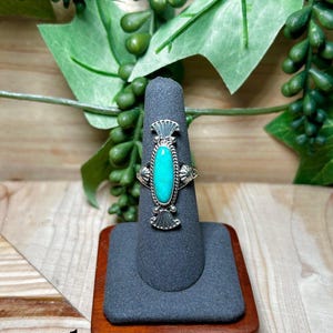 May include: A silver ring featuring an oval turquoise stone set in a twisted wire border. The metalwork includes fan-shaped accents at the top and bottom of the stone setting. The number 1 is printed in black on the lower left side of the image.