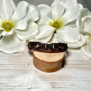 May include: A copper cuff bracelet featuring a repeating pattern of raised diamond shapes and etched triangular details. The metal has a dark, oxidized finish in the recessed areas, contrasting with the polished, metallic copper highlights.