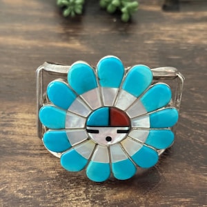 May include: A silver ring featuring a turquoise and mother-of-pearl flower design. The flower's center has a face design with red, blue, and white accents. The ring is set against a wooden surface.