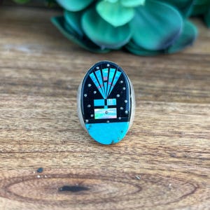 May include: Oval-shaped silver ring with a black inlay featuring a turquoise and multi-colored geometric design. The ring has a turquoise base and is set against a wooden surface.