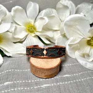 May include: A copper cuff bracelet with a dark, etched design, resting on a small, round wooden stand. The bracelet features a repeating pattern. The background includes white flowers and a gray, textured fabric.