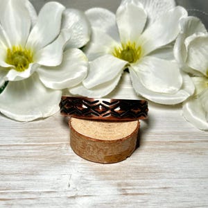 May include: A copper cuff bracelet featuring a repeating geometric pattern of embossed diamonds and horizontal lines. The metal has a polished finish with darkened recessed areas that highlight the intricate texture of the design.