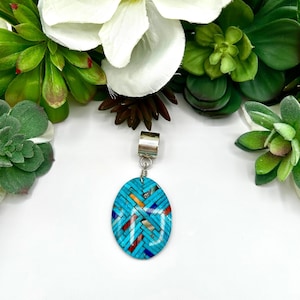May include: An oval pendant featuring a mosaic inlay of turquoise, red, orange, and dark blue stone segments arranged in a woven pattern. The pendant is attached to a polished silver-toned metal bail at the top, creating a secure loop for a chain.