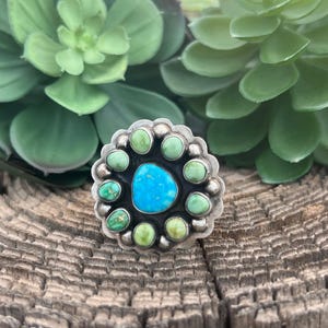 May include: A silver ring with a central turquoise stone surrounded by smaller green stones. The ring has a floral design and is set against a textured wooden surface, with green plants in the background.