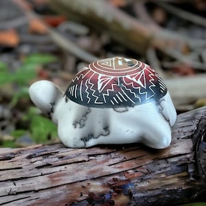 以下が含まれることがあります： クリーム色の体と装飾的な甲羅を持つセラミック製のカメの置物。甲羅は赤、黒、白の幾何学模様です。置物は木の切れ端の上に置かれています。
