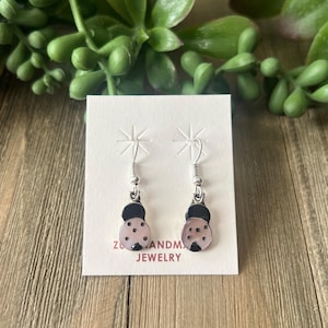 May include: A pair of ladybug earrings on a white card with the text "Z & ANDM JEWELRY". The earrings have a silver hook and a pink body with black spots and a black head. The background includes green foliage and a wooden surface.