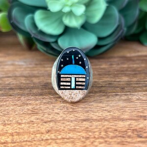 May include: Silver ring with an oval turquoise and black inlay design. The inlay features a stylized building with a blue roof under a starry night sky. The ring is set against a wooden surface with a succulent plant in the background.