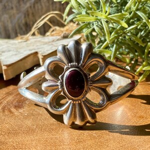 Puede incluir: Una pulsera de plata con una piedra ovalada de color rojo oscuro en el centro, engastada en un marco de metal pulido. El diseño consta de bucles de plata simétricos en forma de pétalos que irradian desde el centro, creando una forma floral.