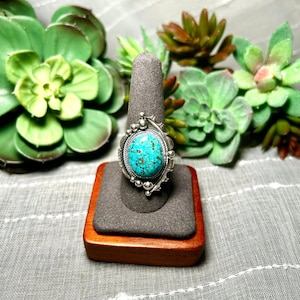 May include: A silver ring featuring an oval-shaped turquoise stone with dark brown matrix patterns. The stone is set in a serrated bezel, surrounded by a twisted wire border and decorative silver spheres along the edges of the metal setting.