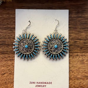 May include: A pair of Zuni handmade silver earrings. Each earring features a central turquoise stone surrounded by a circle of smaller stones and silver accents. The design is further embellished with radiating turquoise rods. The earrings are displayed on a white card.