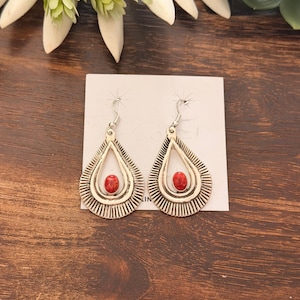 May include: A pair of silver-toned teardrop earrings with a red stone center. The earrings have a detailed, ridged design around the edges and are displayed on a white card. The background is a dark wood surface.