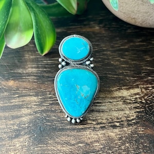 May include: A silver ring featuring two bright blue turquoise stones set in a vertical arrangement. The top stone is circular, while the bottom stone is a larger teardrop shape. Both stones are framed by twisted silver wire and small silver beads.
