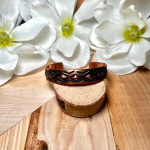 May include: A copper cuff bracelet with a geometric pattern in black. The bracelet is displayed on a small wooden stand, with white flowers in the background. The bracelet has a width of approximately 0.75 inches.